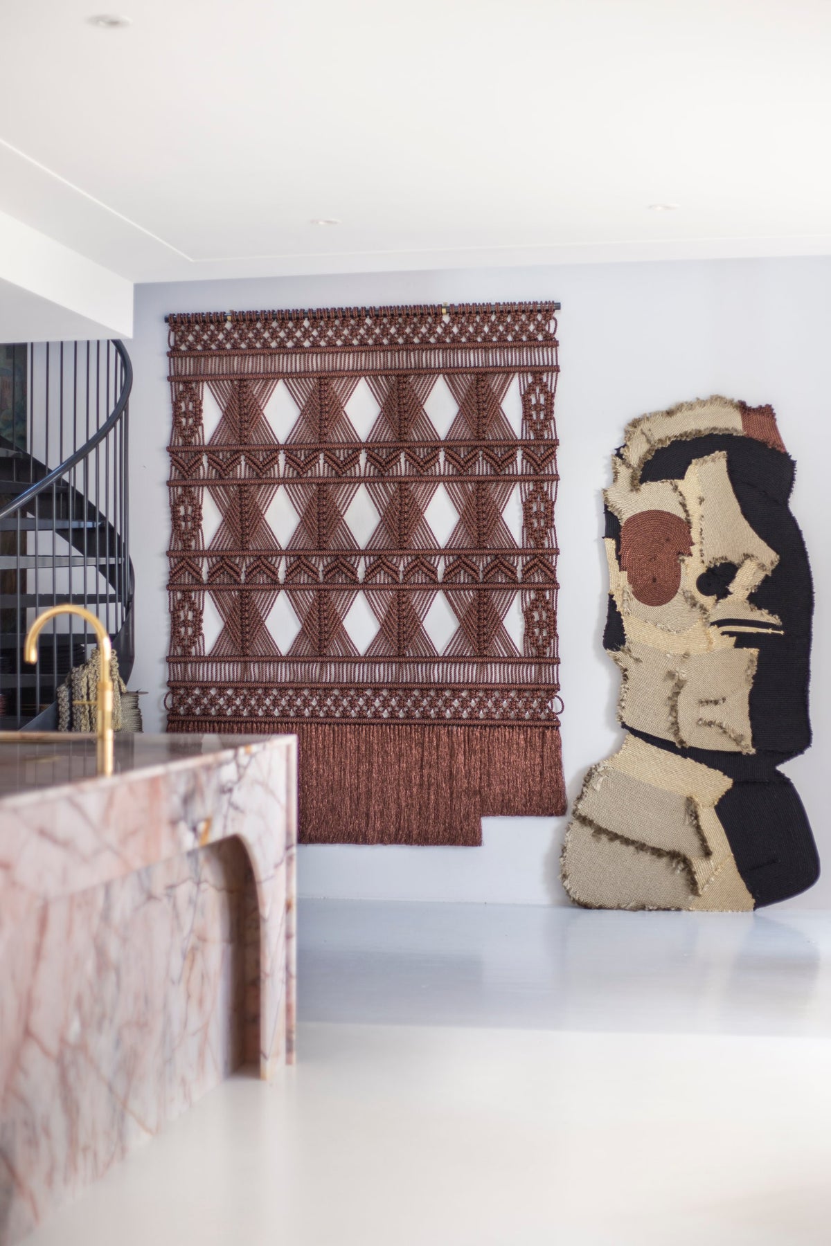 Brown metallic macramé wall hanging beside a fiber art figure, seen past a pink marble kitchen island